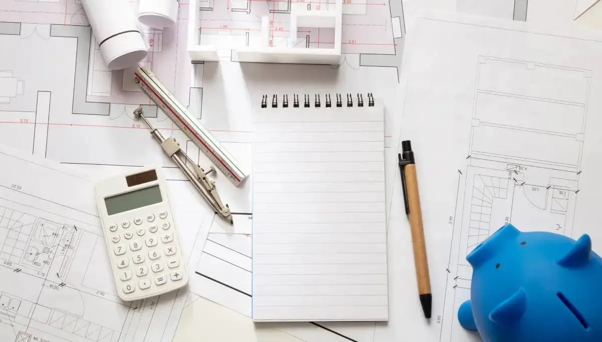 a notepad, pencils, calculator, and blue piggy bank on top of a remodeling blueprint as the contractor creates a cost estimate
