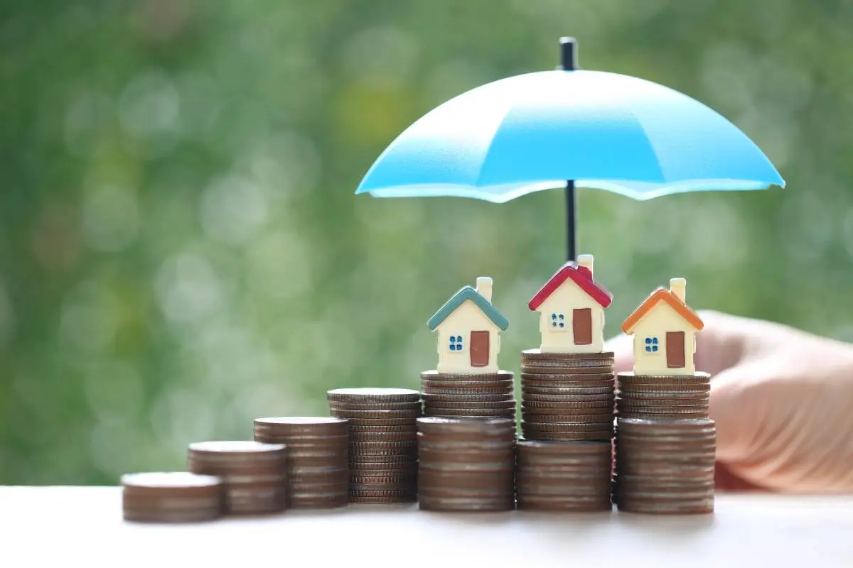 a hand holding blue umbrella over house figurines on a ramp of stacked coins