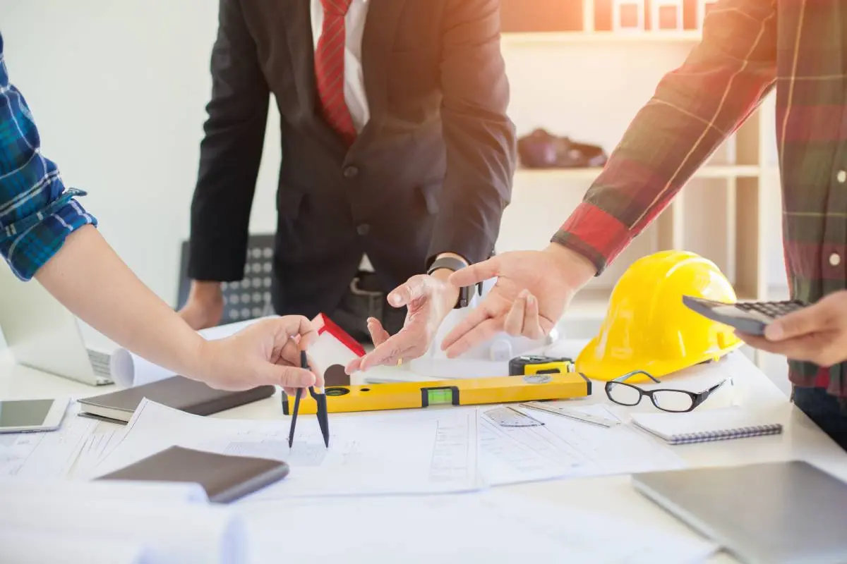 a man in a suit with two men in plaid button-shirts reviewing blueprints on a desk with a hardhat, level, tape measure, and glasses