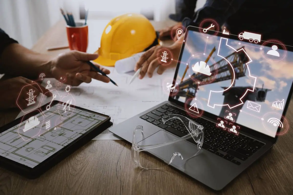 two contractors at a table with pencils in their hands going over blueprints with a hard hat to one side of them and a laptop and tablet to the other side with icons from contractor software floating in front