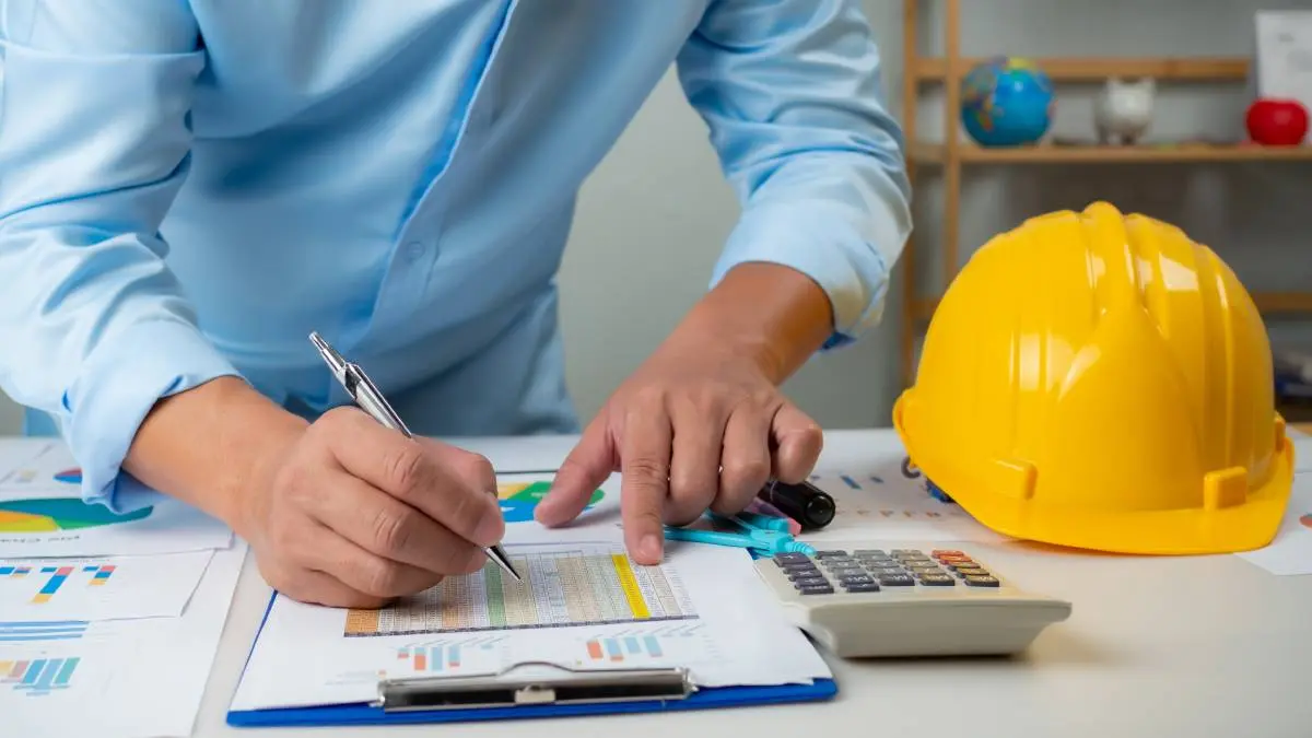 a man in a blue button-down shirt writing on a chart on a clipboard with a calculator and yellow hard hat next to him on his desk
