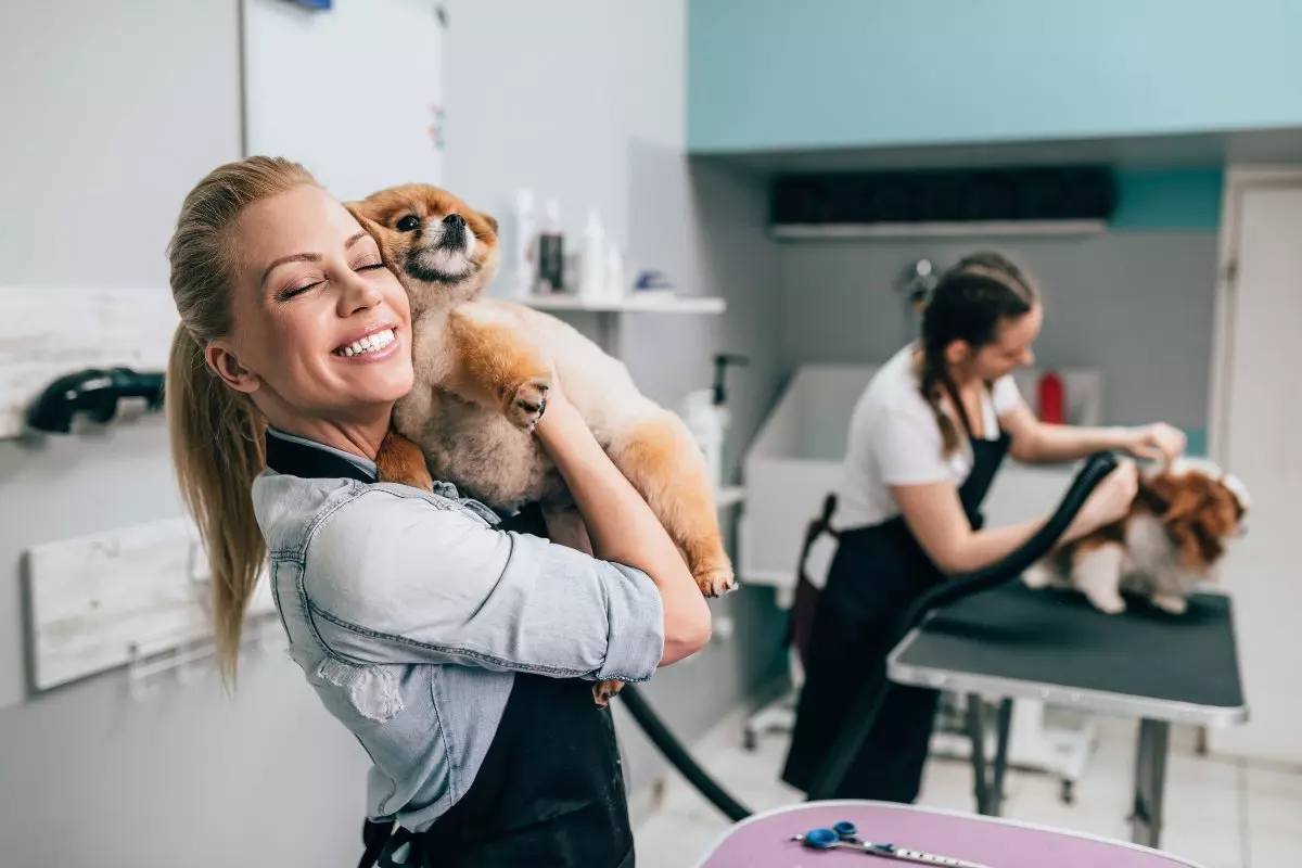 Groomer hugging dog with another groomer cutting dog's hair in background 