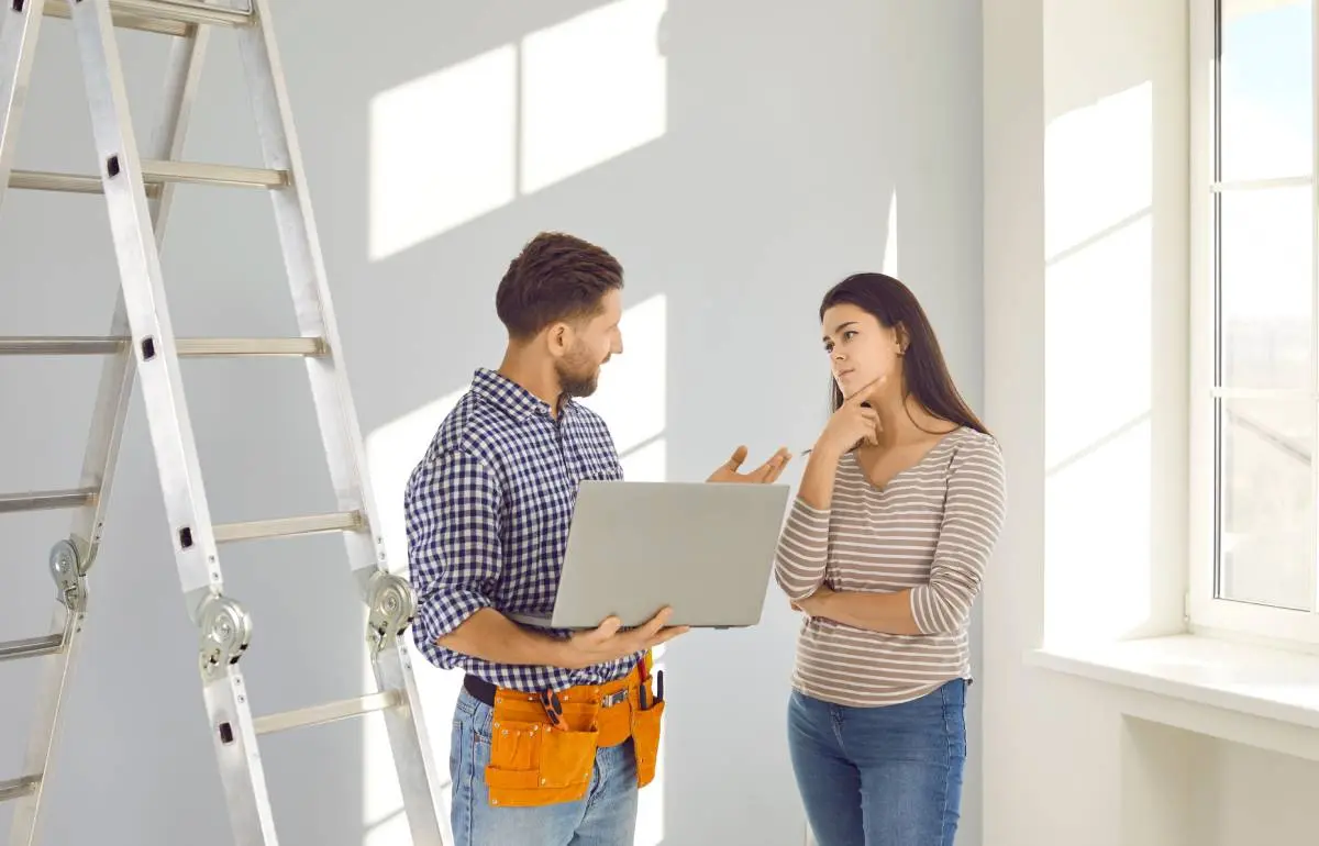 a contractor wearing a tool belt and holding a laptop explaining a bid to a homeowner in a striped shirt in an room empty but for a ladder