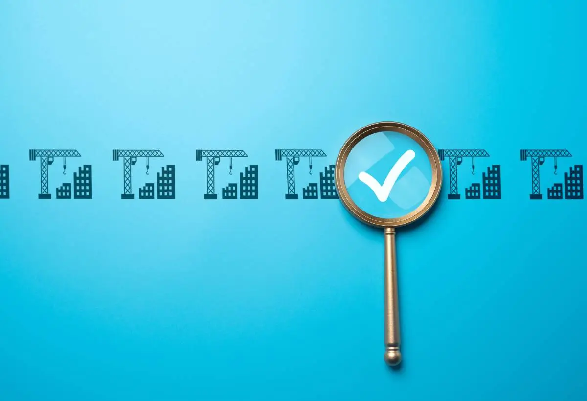 a gold magnifying glass highlighting a white checkmark surrounded by icons of a crane constructing two buildings against a blue background