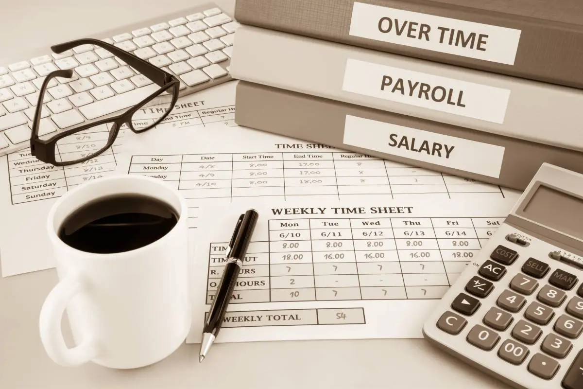 calculator, cup of coffee, keyboard, glasses, printed-out weekly time sheets, and binders marked overtime, payroll, and salary on a desk in shades of gray