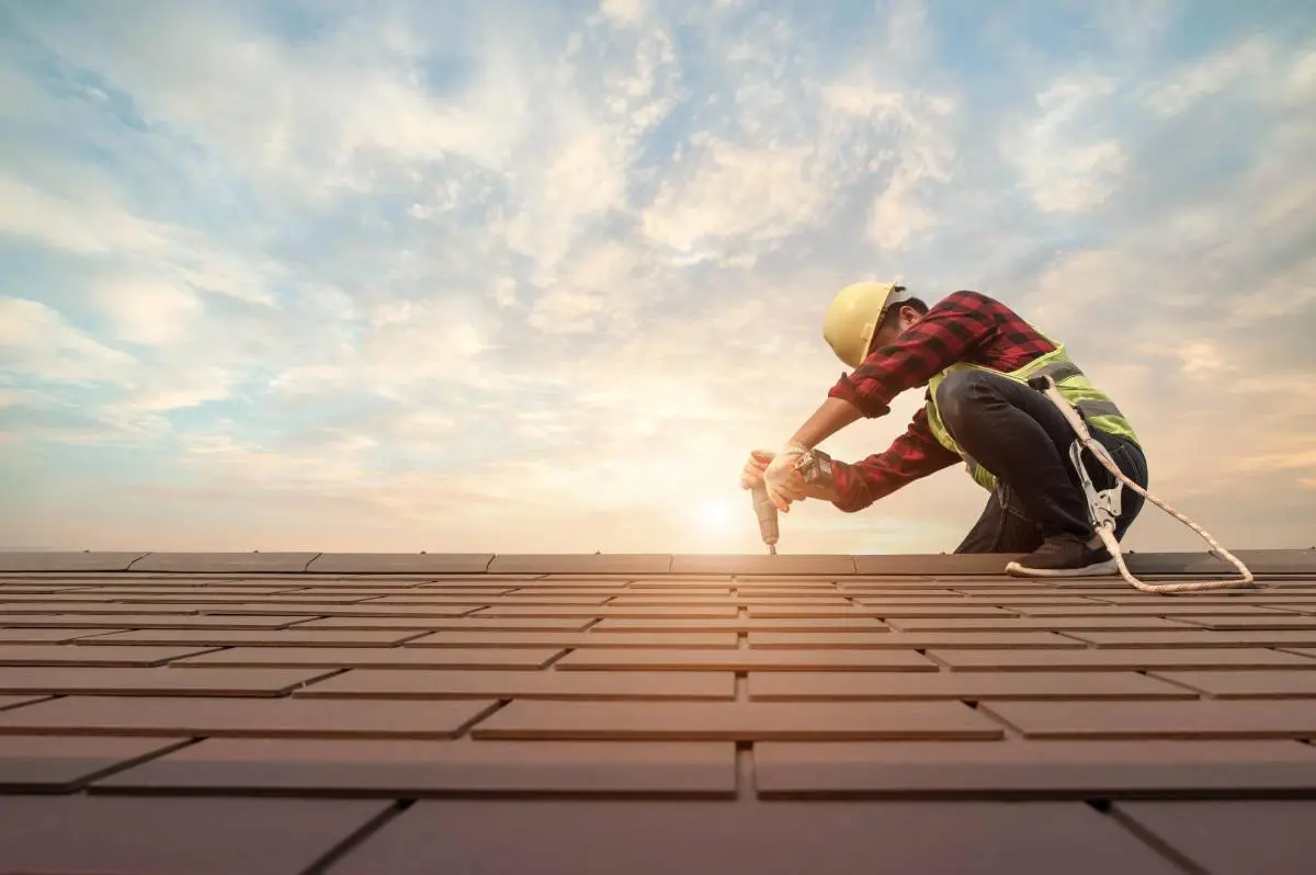 a roofing contractor wearing a red and black plaid shirt and yellow hardhat using a power drill on a roofing project with a sunset behind him