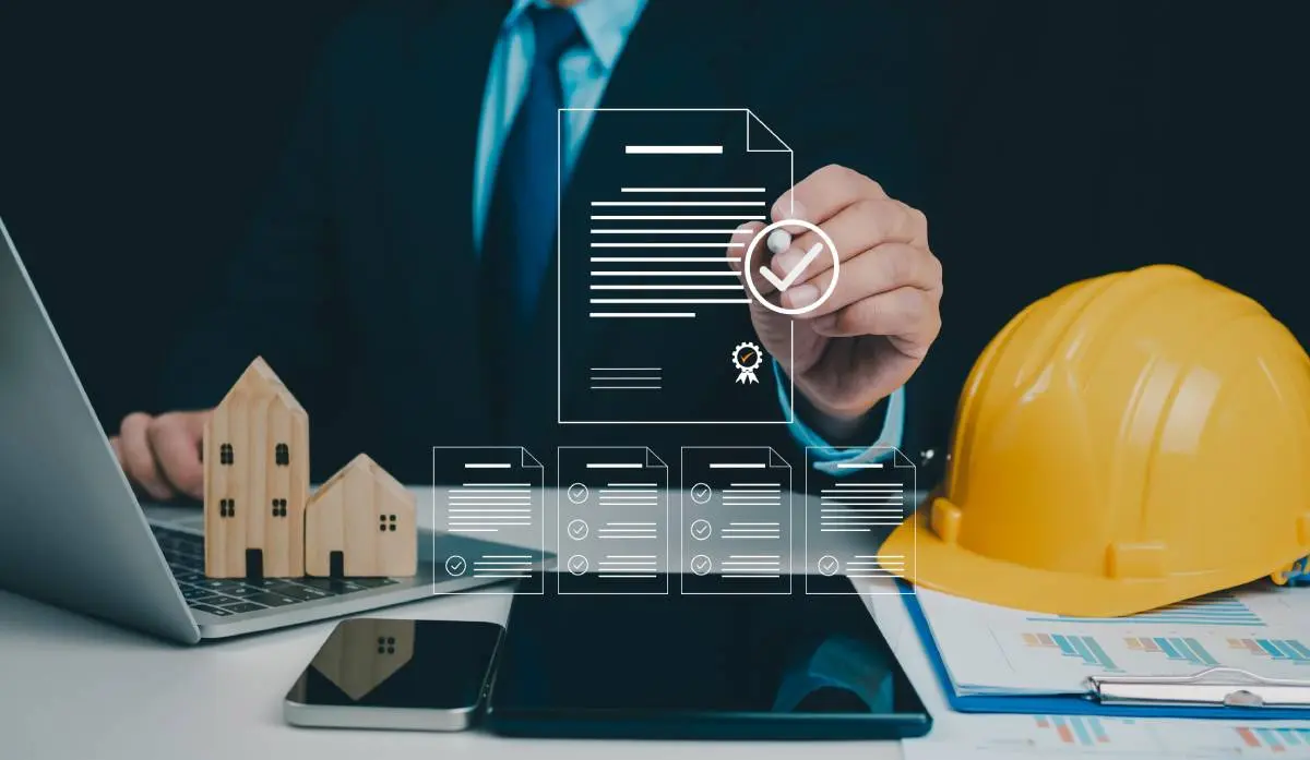 a yellow hardhat, laptop, cellphone, and tablet spread out in front of a man wearing a suit and tie as he fills out digital applications for a roofing contractor license 