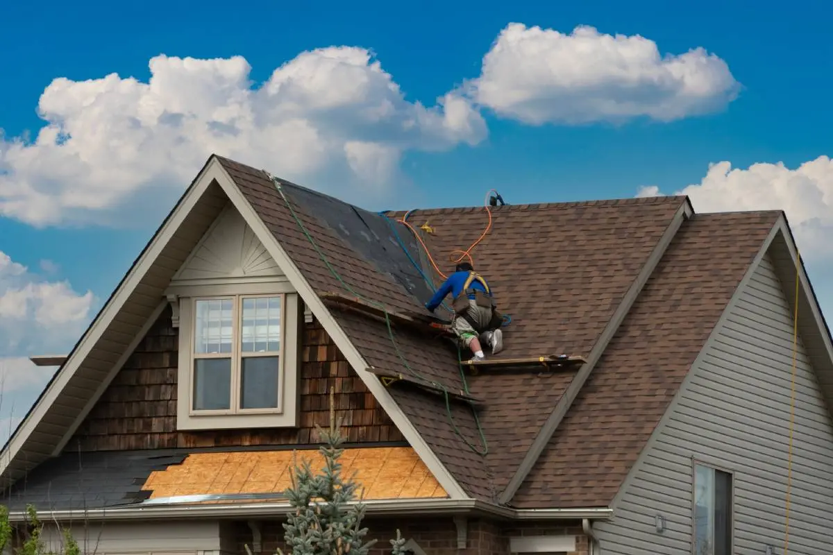 a roofing contractor wearing a blue shirt working on a roofing project with wires and other tools as the sky and clouds are behind him