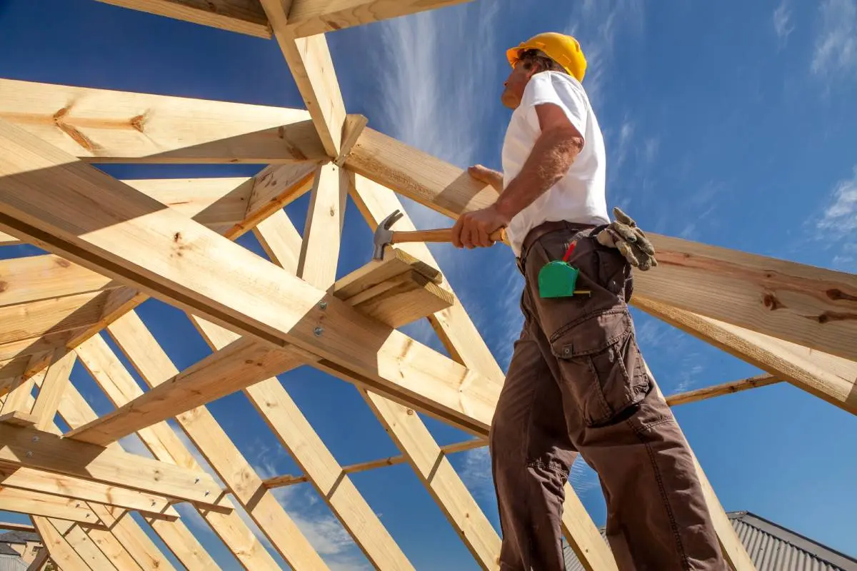 a roofing contractor holding a hammer and working on foundational wooden beams with a blue sky behind him
