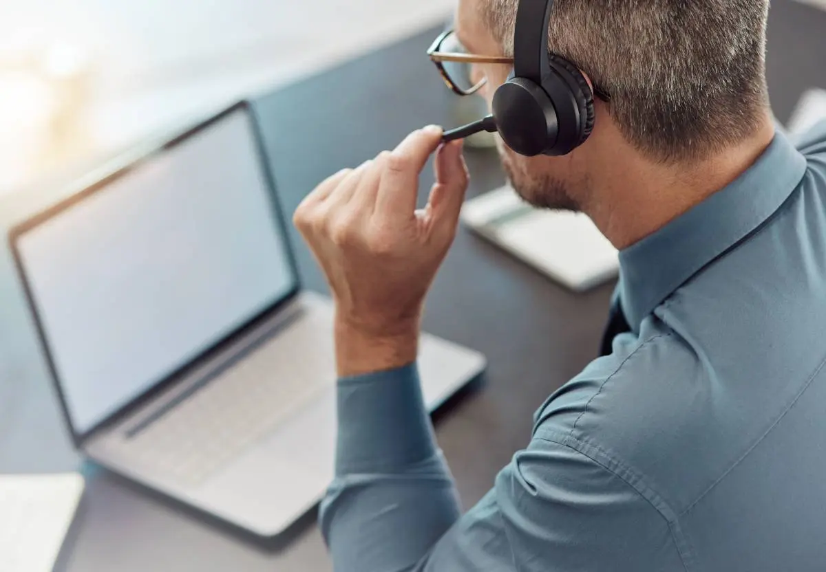 a man in a blue button down shirt on a sales call with a headset as he sits at a desk with his laptop