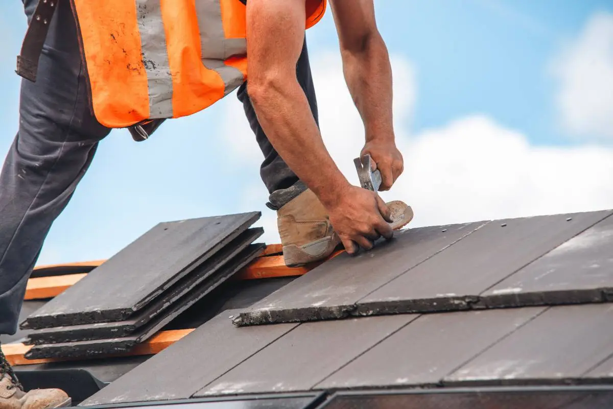 a roofing contractor in an orange vest hammering in nails to tiles on a roof during the summer months