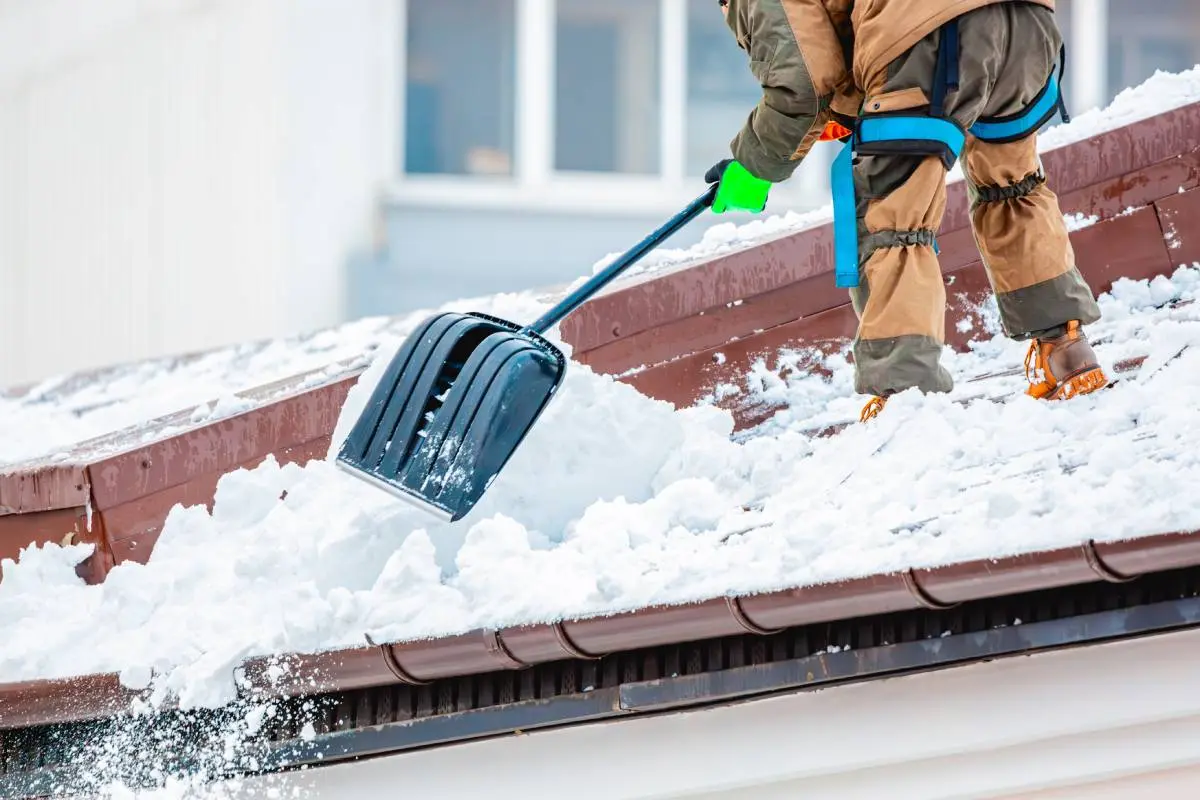 a roofing contractor in winter gear performing seasonal maintenance by shoveling snow off a roof