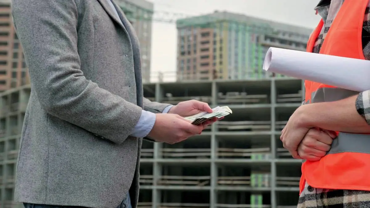 a contractor in a gray blazer and blue button-down shirt counting cash to payout his subcontractor wearing a plaid shirt and orange safety vest