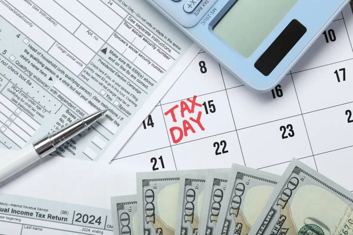 a calendar with "tax day" written in red marker with hundred dollar bills, a calculator, and an income tax return form on top of it